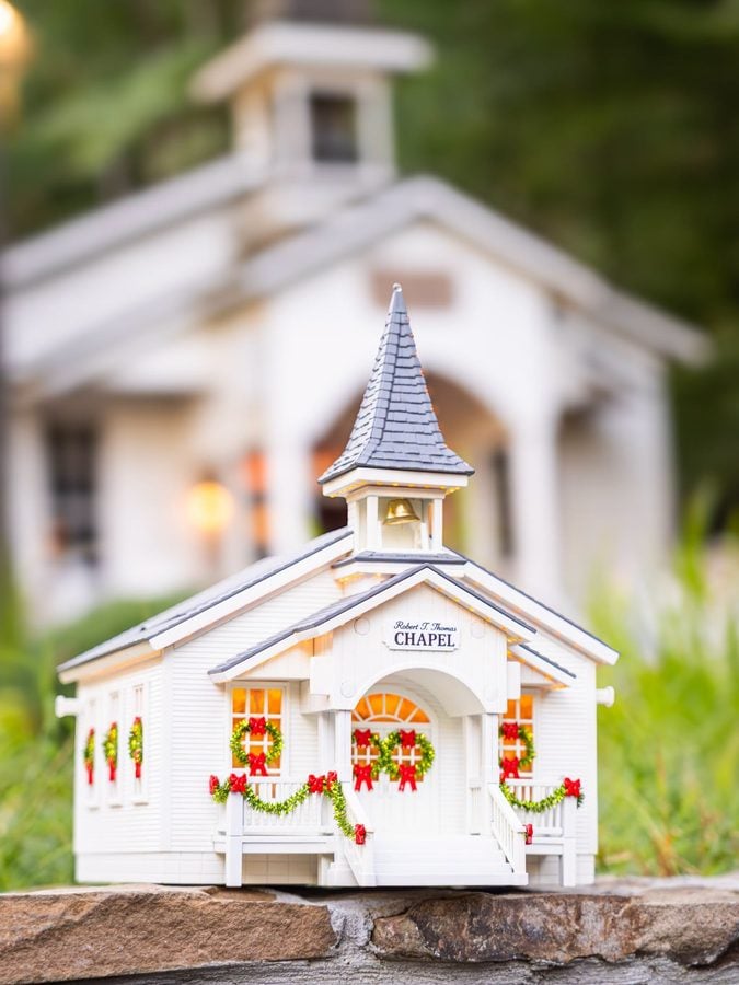 Small white miniature chapel with a blue gray shingled roof decorative wreaths and red ribbons on the exterior placed on a stone surface with a blurred house backdrop