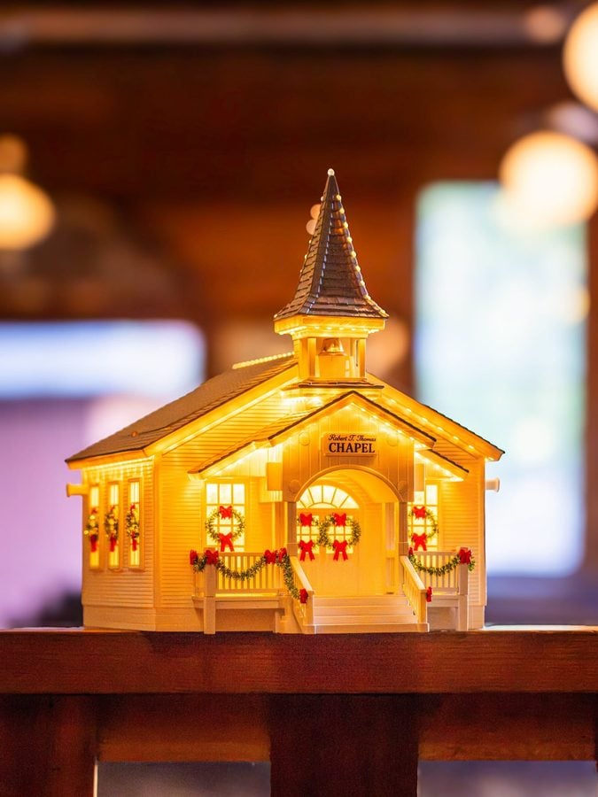 Lit miniature wooden chapel with a tall pointed steeple warm yellow lights and festive wreaths and red bows on the porch placed on a wooden shelf