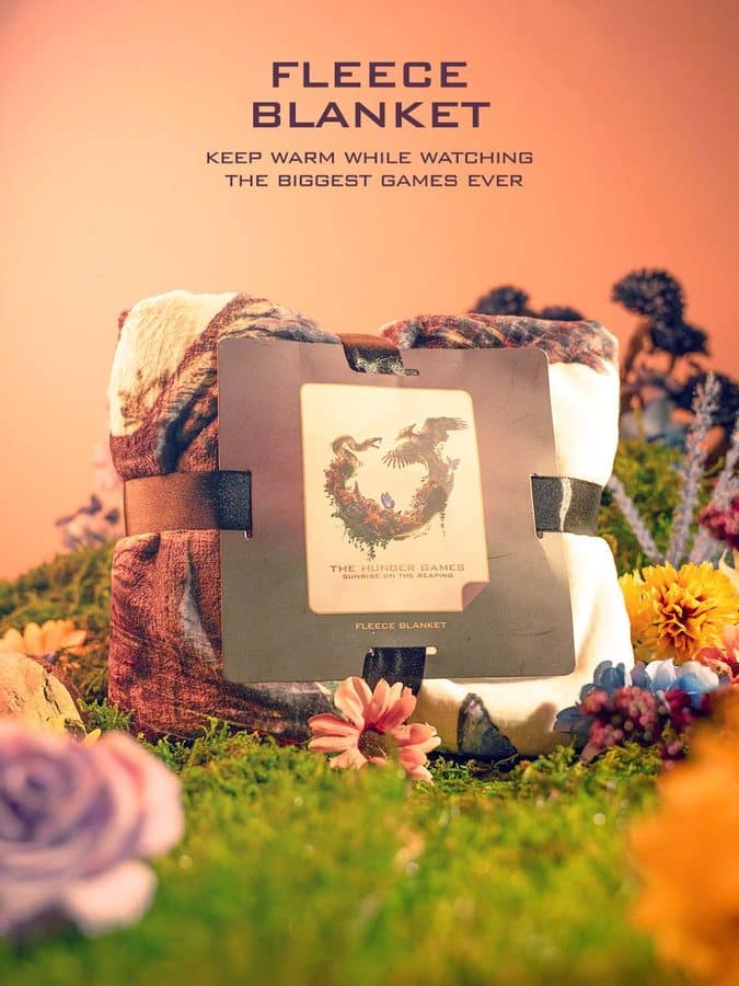 Fleece blanket folded on a pedestal with a Hunger Games label placed in a colorful garden scene at sunset promoting warmth