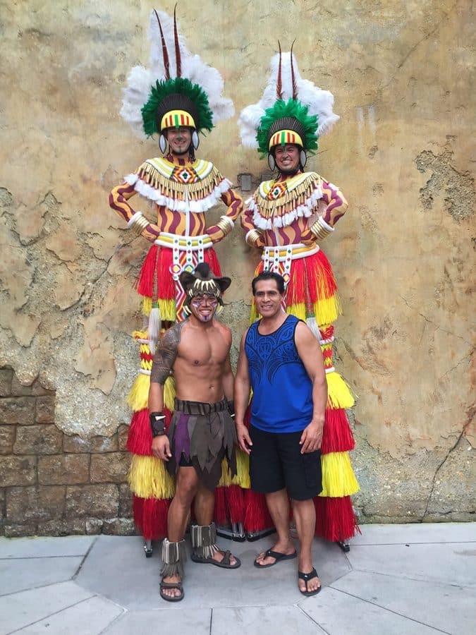 Two large feathered dancers in colorful traditional costumes pose with two men against a textured stone wall exterior