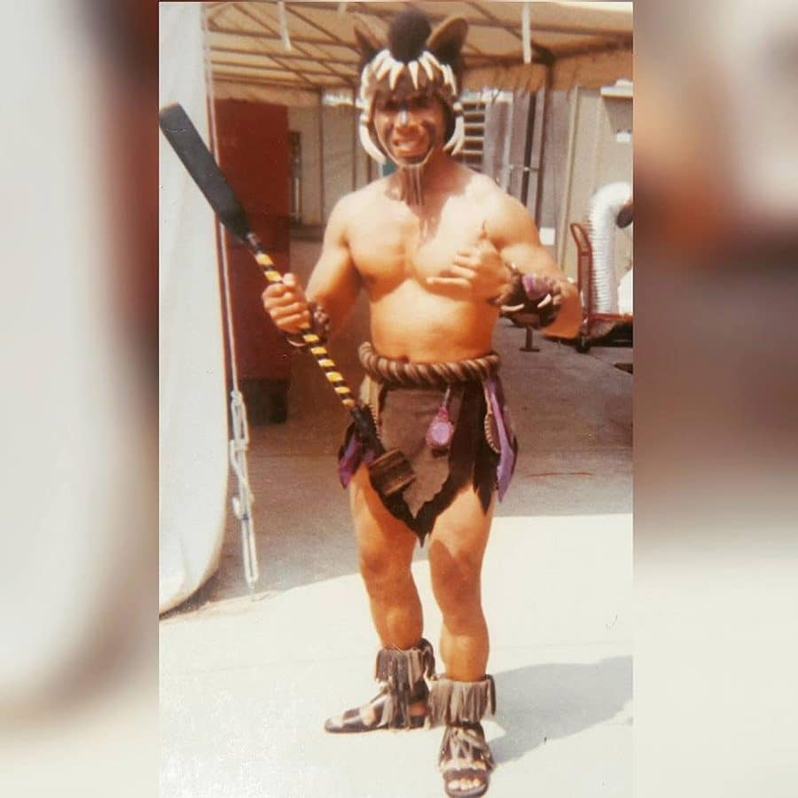 Shirtless man in a tribal warrior costume with a tiger head headdress holding a spear outdoors in a festival like setting