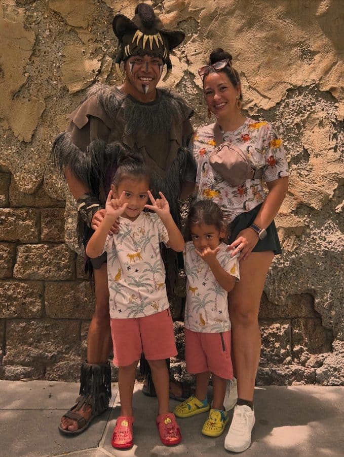 Family posing with a performer in a fur trimmed tribal costume and bone headdress beside a smiling woman in a floral shirt two kids in matching jungle print outfits in front against a rough stone wall backdrop