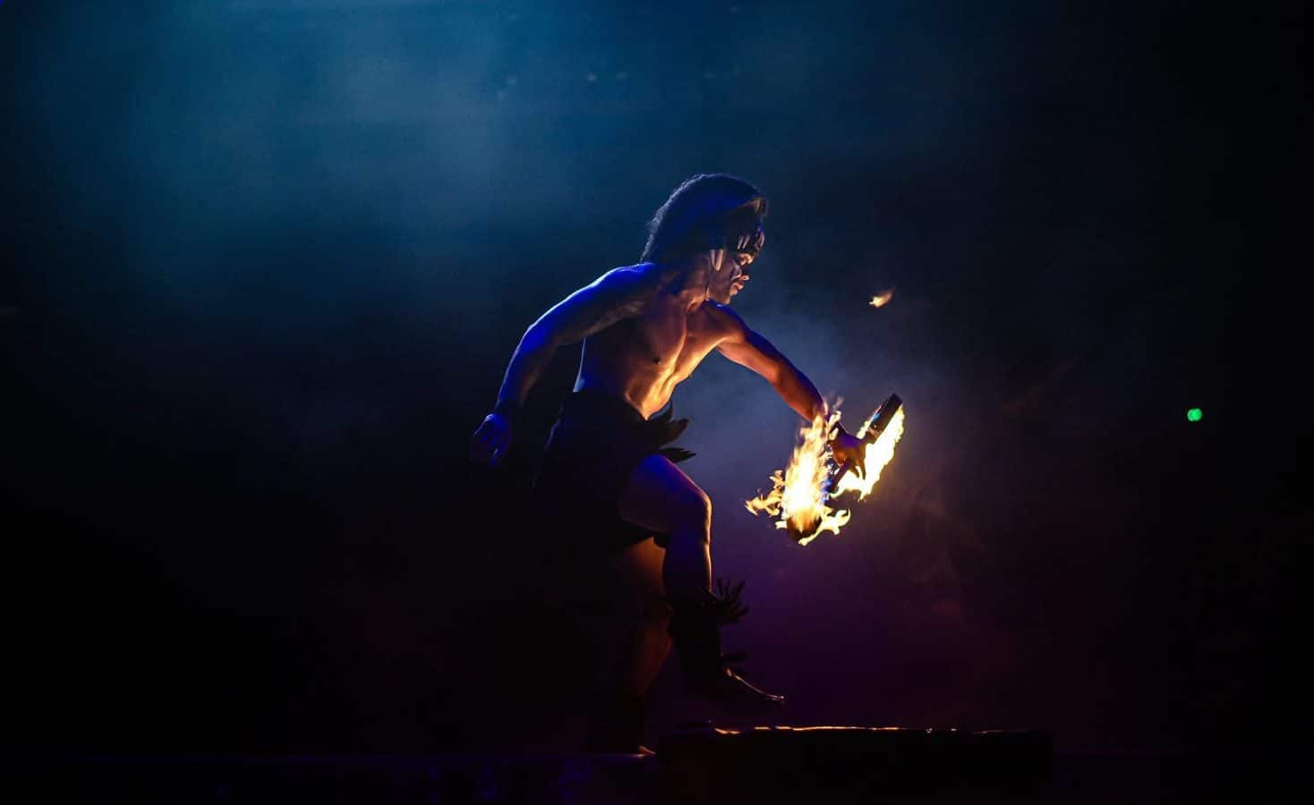 A shirtless fire performer spinning flaming poi against a dark night backdrop
