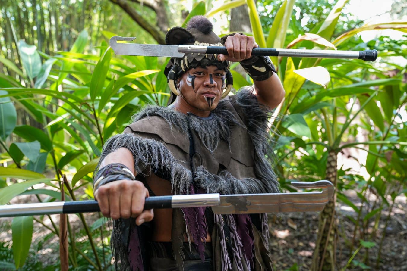 Person in tribal costume with face paint gripping a long spear in a lush outdoor setting jungle like garden