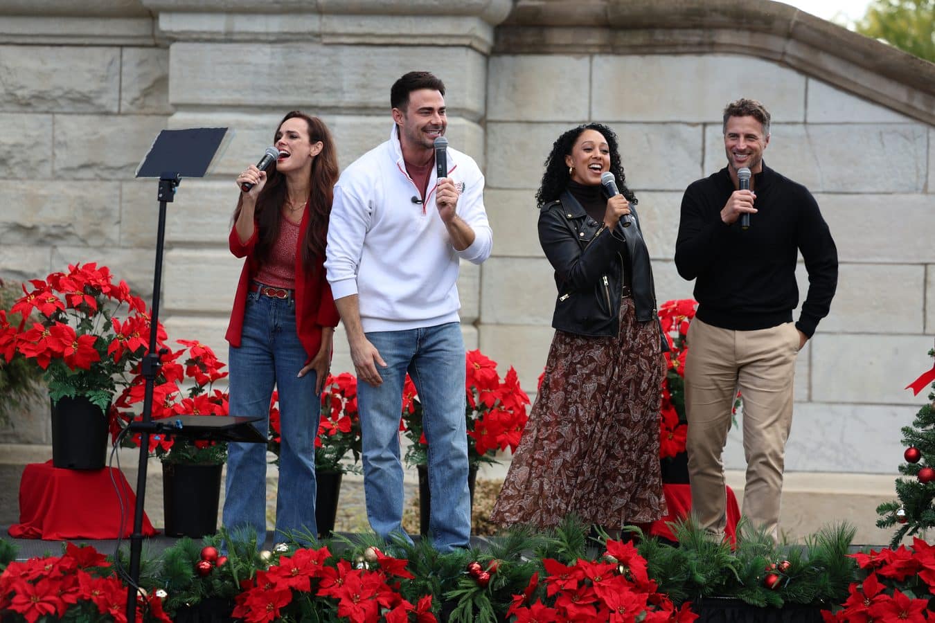 Four singers perform on an outdoor stage each holding a microphone with bright red poinsettias and holiday greenery around them