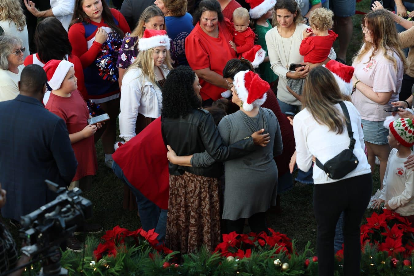 Crowd in Santa hats and red clothing gathered in a circle on grass with poinsettias along the edge