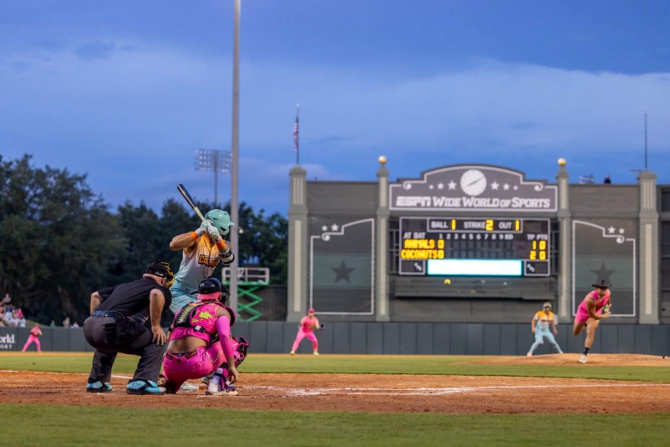 Batter in a teal helmet swings at a pitch at home plate with a pink clad catcher and umpire nearby and a large scoreboard in the background at a stadium