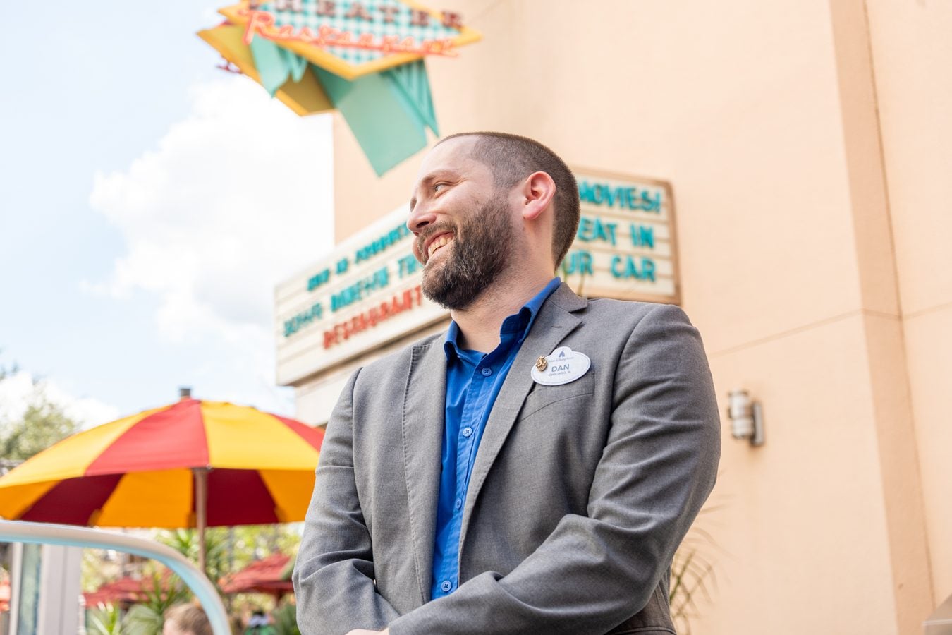 Smiling bearded man in a gray blazer and blue shirt with a name badge outdoors near colorful umbrellas and a sign in the background