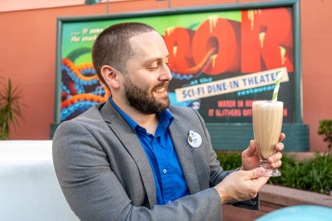 Smiling man in a gray blazer and blue shirt holding a tall milkshake with a green and white striped straw outdoors with a colorful poster behind him