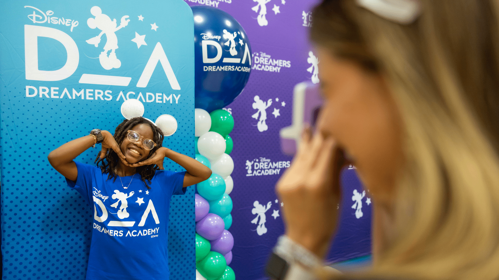 Smiling girl in a blue Dreamers Academy shirt poses with hands under her chin in front of a Disney backdrop and balloon column photographer blurred in foreground