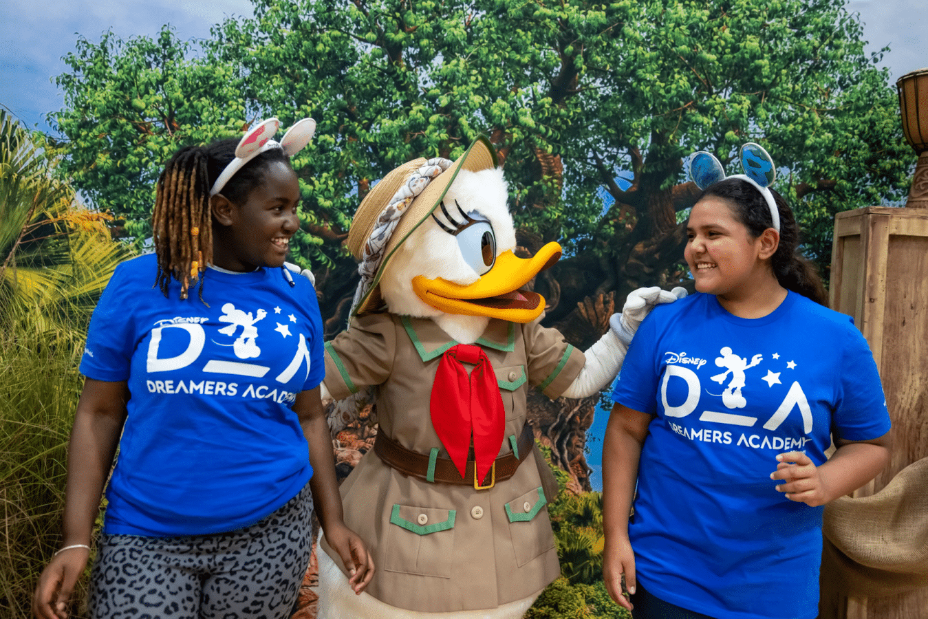 Two girls in blue Dreamers Academy shirts pose and smile with a costumed Donald Duck in a safari outfit