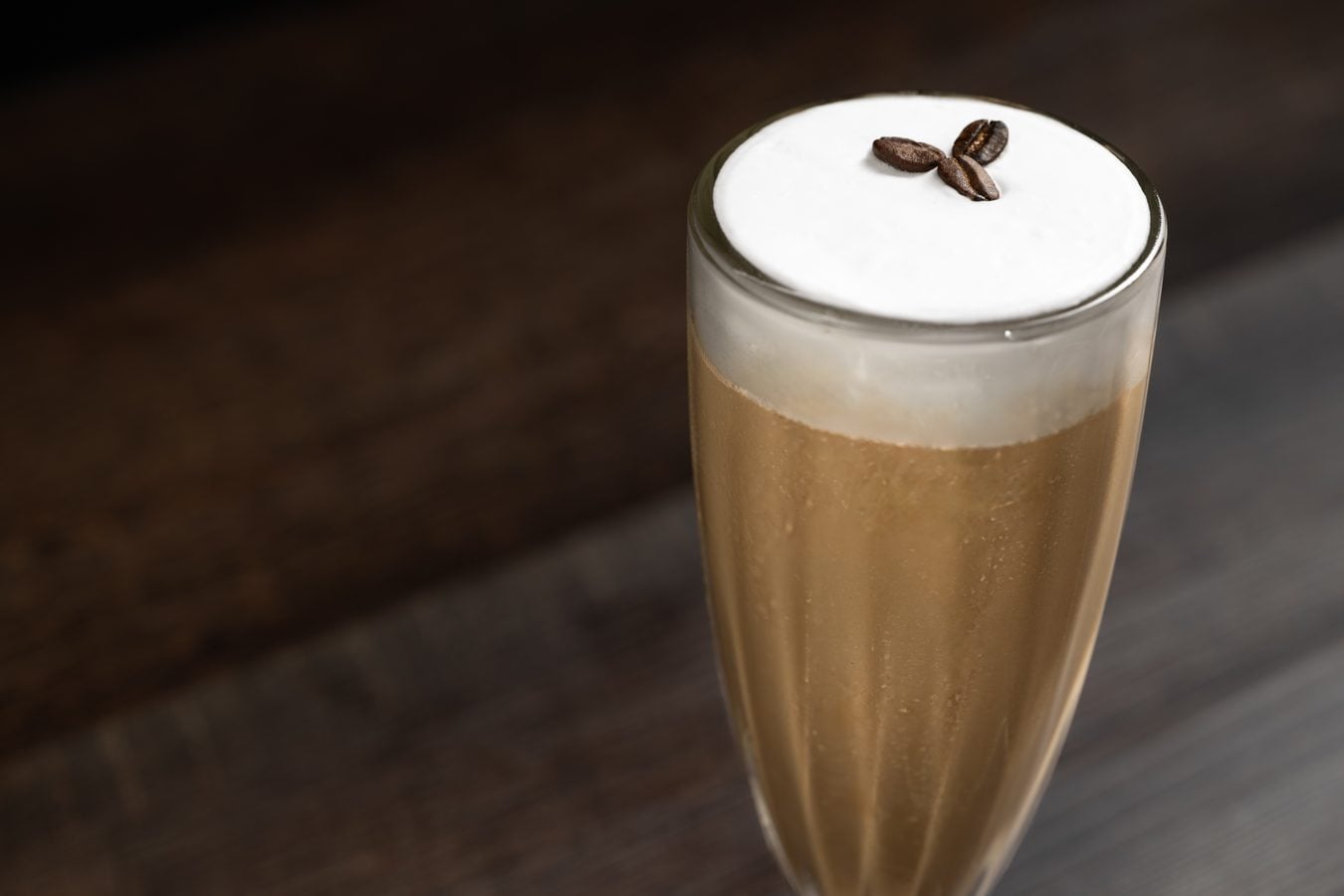 Close up of a tall glass latte with a thick white foam top and two coffee beans resting on it