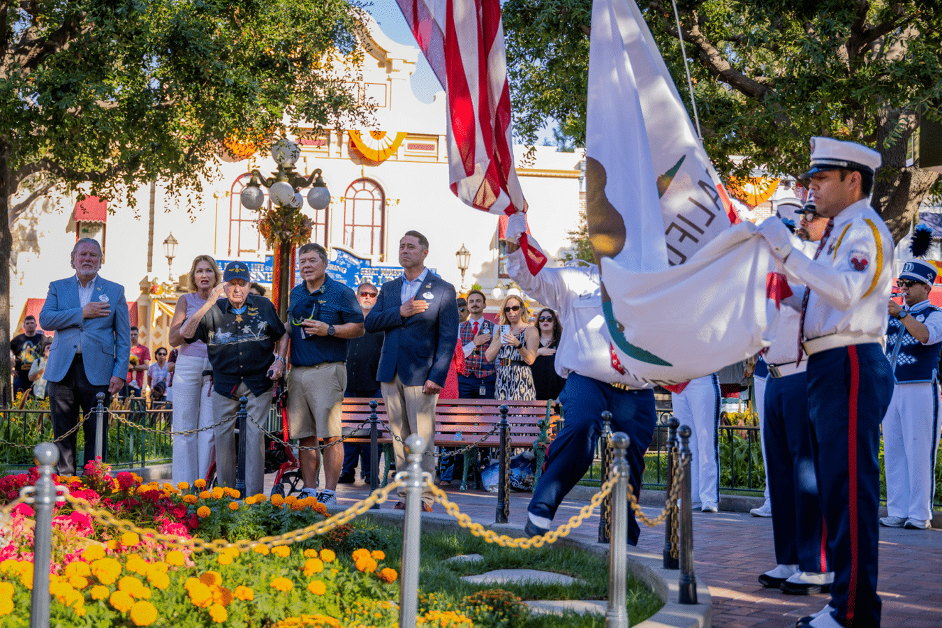 Civilians and service members stand with hands over their hearts during a patriotic ceremony in a sunny plaza with a colorful flower bed in the foreground and flag bearers nearby
