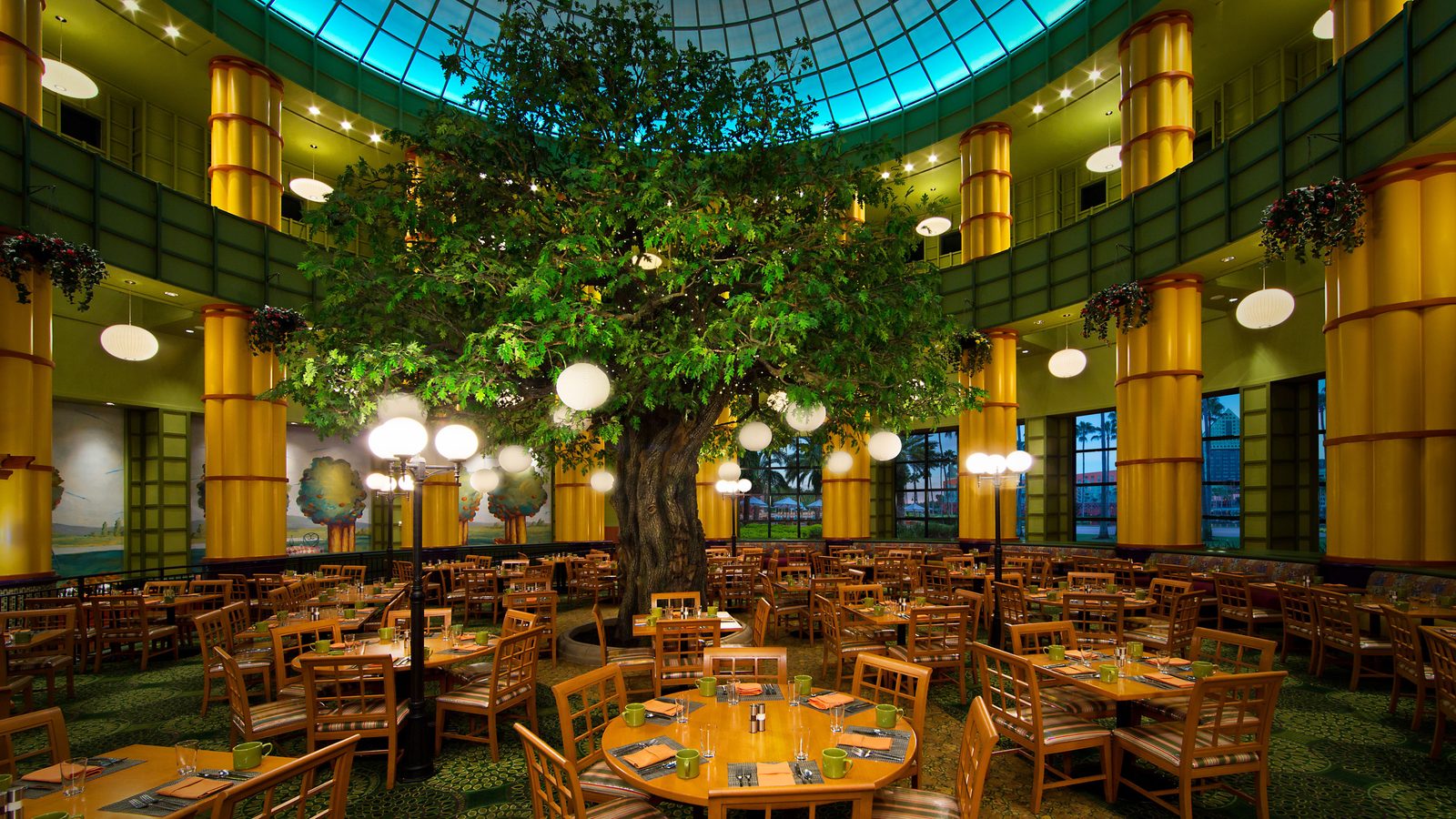 Elegant indoor dining hall with a large tree at center surrounded by round tables and warm globe lights in a tropical themed atrium