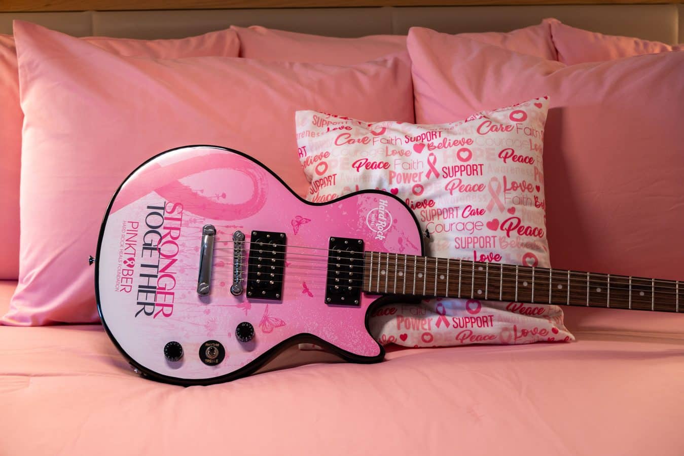 Pink electric guitar resting on a pink bed with a heart themed pillow beside it