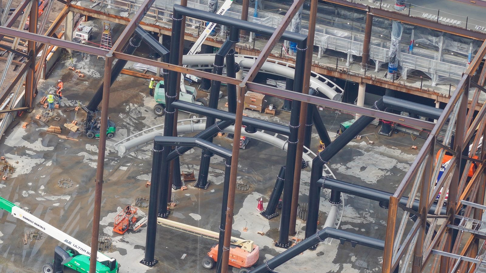 A construction site with dark steel beams forming a grid workers in safety vests and heavy equipment on a wet concrete ground viewed from above