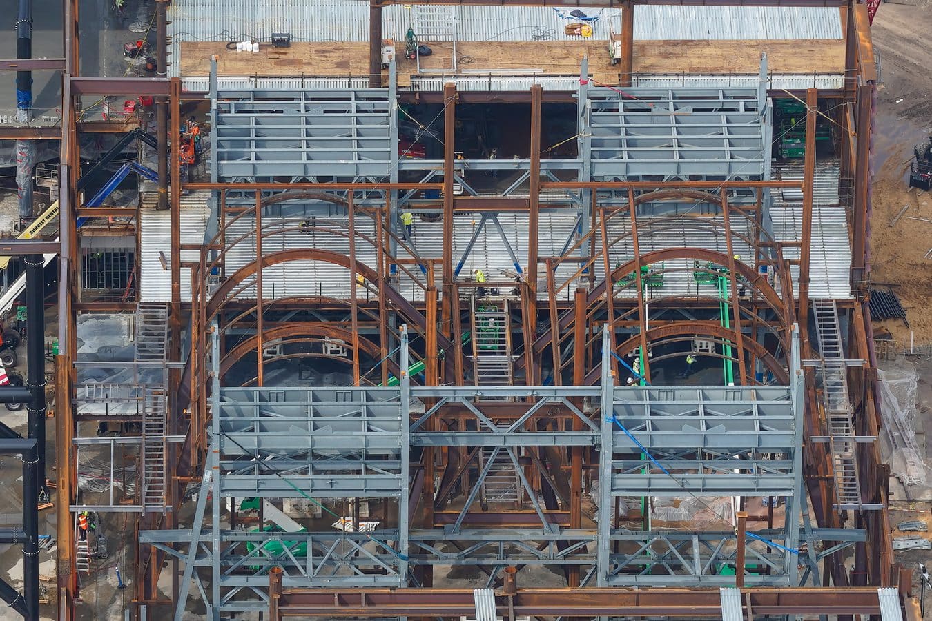 Aerial view of a large steel framed construction site with gray decking rust red beams ladders and workers in safety vests