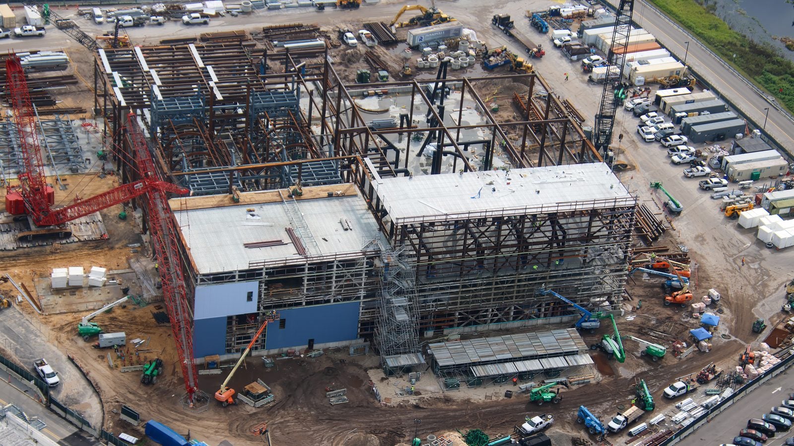 Aerial view of a large construction site with a steel framed building cranes and construction vehicles scattered around a muddy yard