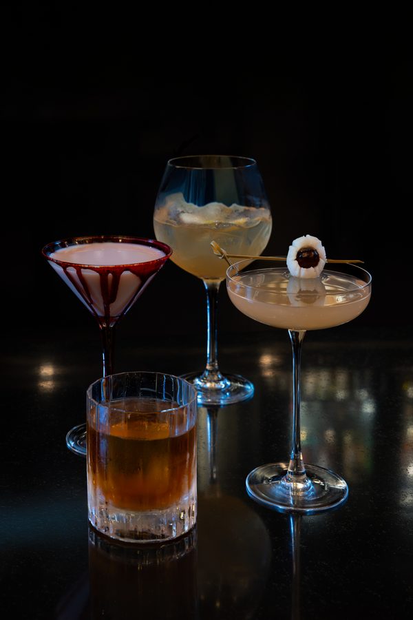 Three cocktails on a dark bar pink drink in a sugar rimmed coupe on the left a pale yellow ice filled beverage in a large stemmed glass center and a pale cocktail with an olive garnish on a skewer in a coupe on the right