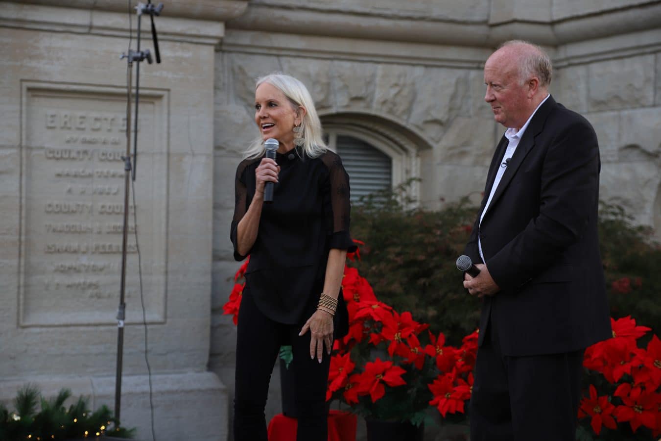 Two speakers on an outdoor stage a woman with a handheld microphone and a man in a suit red poinsettias behind them