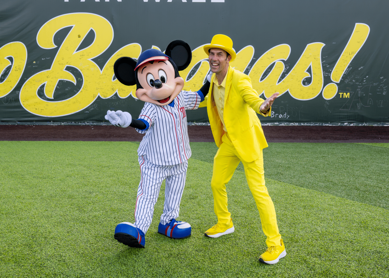 Mickey Mouse mascot and a man in a bright yellow suit posing with arms outstretched on a baseball field with a large yellow banner in the background