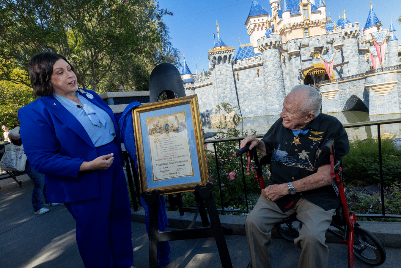 Woman in a blue suit presents a framed certificate to an elderly man in a wheelchair with a castle backdrop and garden