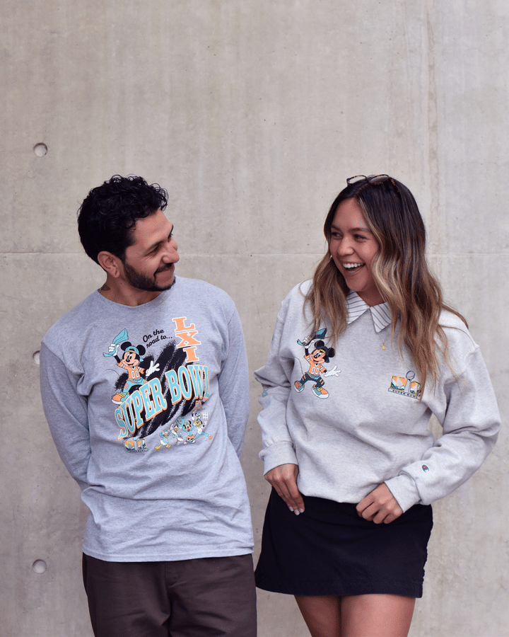 Two people in gray Mickey Mouse Super Bowl sweatshirts smile at each other while standing against a concrete wall
