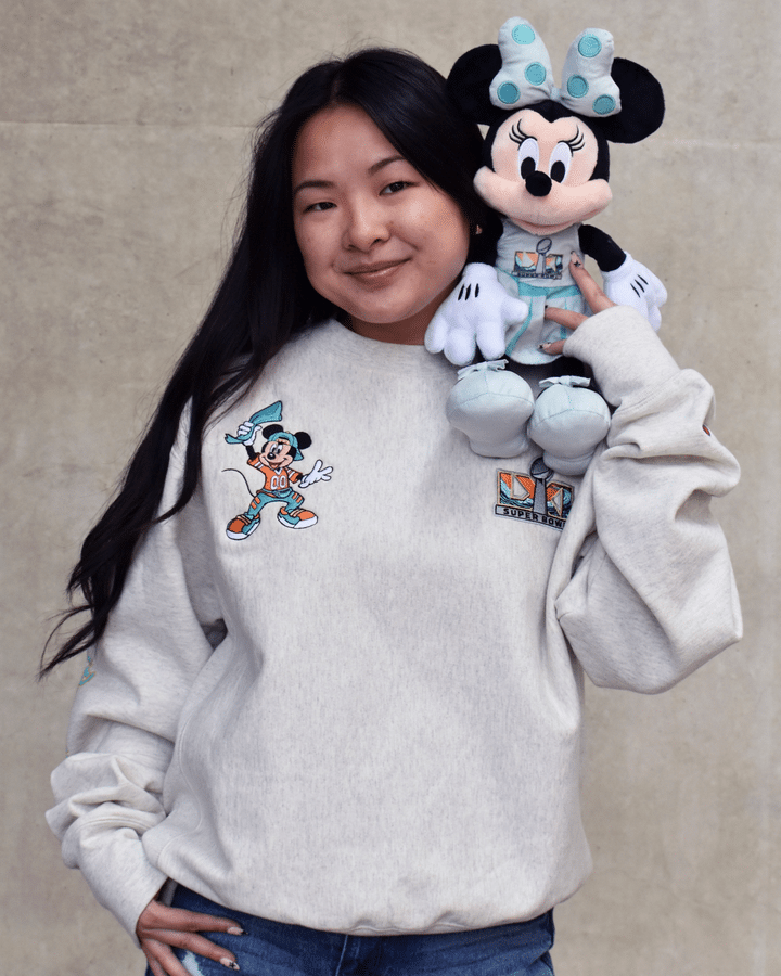 Young woman in a gray sweatshirt smiles while holding a Minnie Mouse plush beside her head against a concrete backdrop