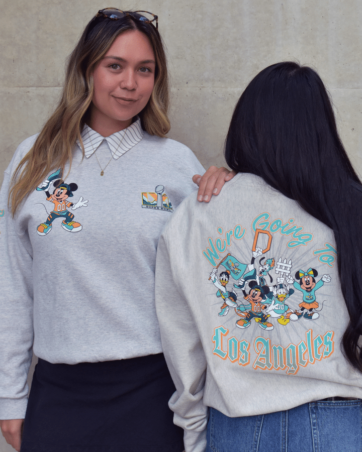 Two young women stand against a concrete wall the left woman faces the camera wearing a gray sweatshirt with a Mickey Mouse graphic and sunglasses on her head while the right woman shows the back of her sweatshirt with a colorful Los Angeles design