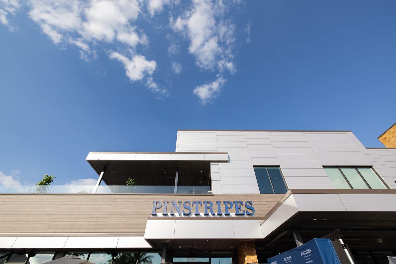 Modern commercial building exterior with a beige panel facade and glass balcony PINSTRIPES sign above the entrance against a blue sky