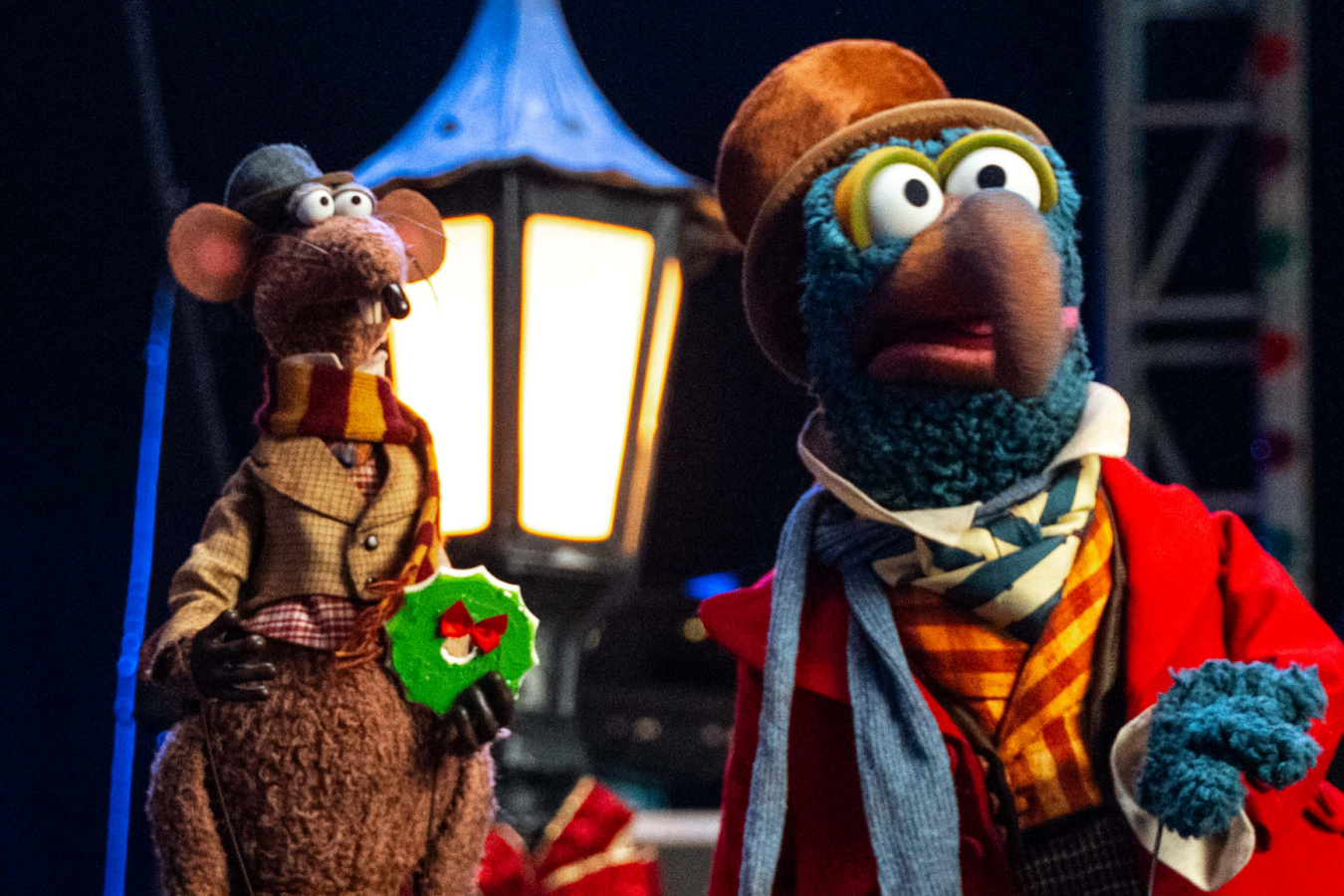 Two felt puppet characters in costumes on a stage one holding a green wreath under a warm lantern light