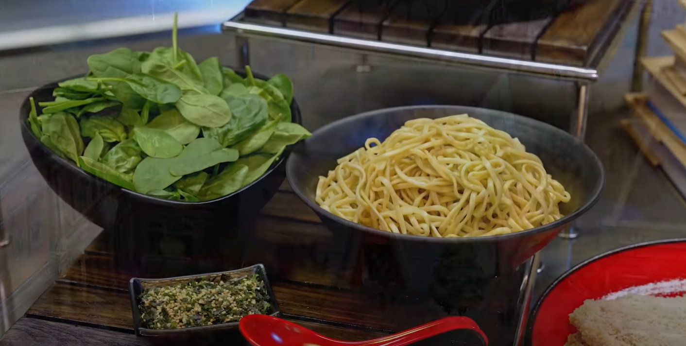 Bowl of fresh spinach next to a large bowl of cooked noodles on a buffet counter