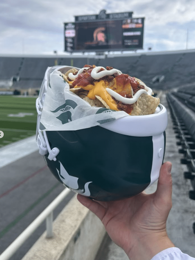 Hand holding a helmet shaped bowl of loaded nachos with cheese at a football stadium with a large scoreboard in the background