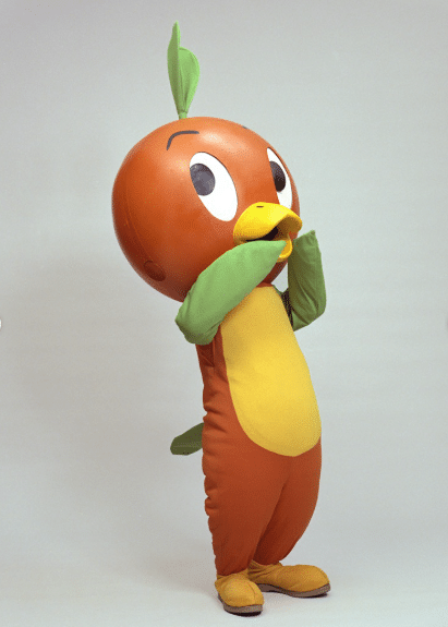 Orange bird mascot with a yellow belly and green wings standing with a leaf on its head and hands near its beak in a playful pose