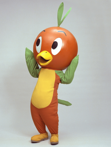 Orange bird mascot standing with arms raised yellow belly green leaf like headpiece and wings posing for a photo