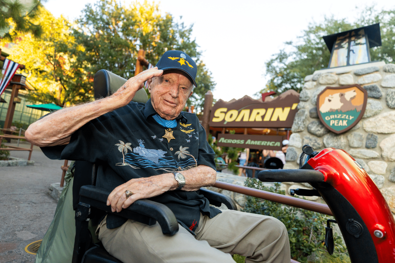Elderly man in a wheelchair salutes with his right hand at an outdoor attraction behind him with a SORIN Across America sign and stone marker nearby wearing a cap and medal necklace