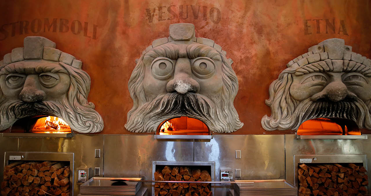 Three carved stone bearded faces above wood fired ovens flames glowing inside and a warm orange wall behind them