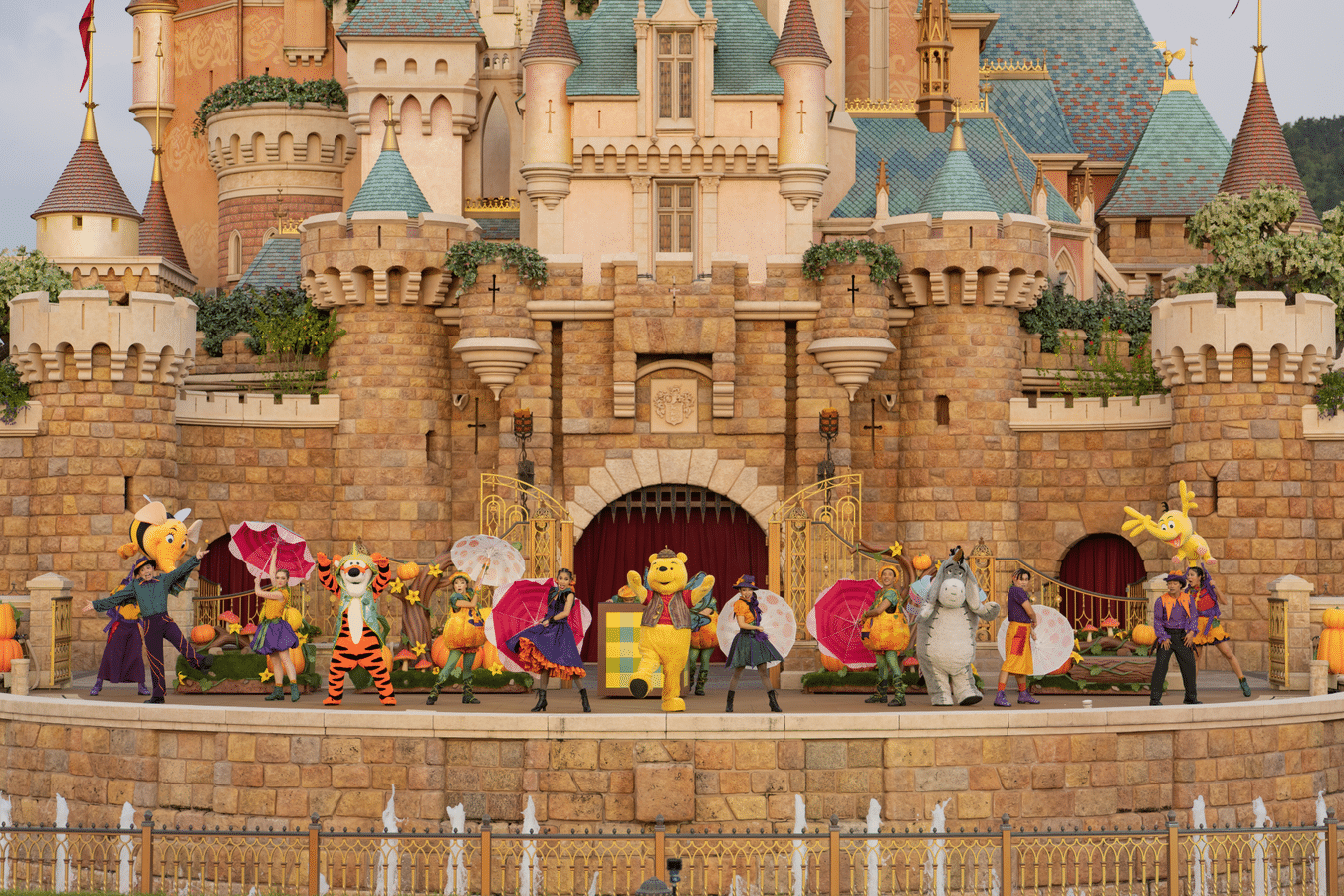 WinniethePoohHongKong Fantasy Land News Colorful parade performers and mascots dancing on a stage in front of a castle with decorative umbrellas and festive costumes