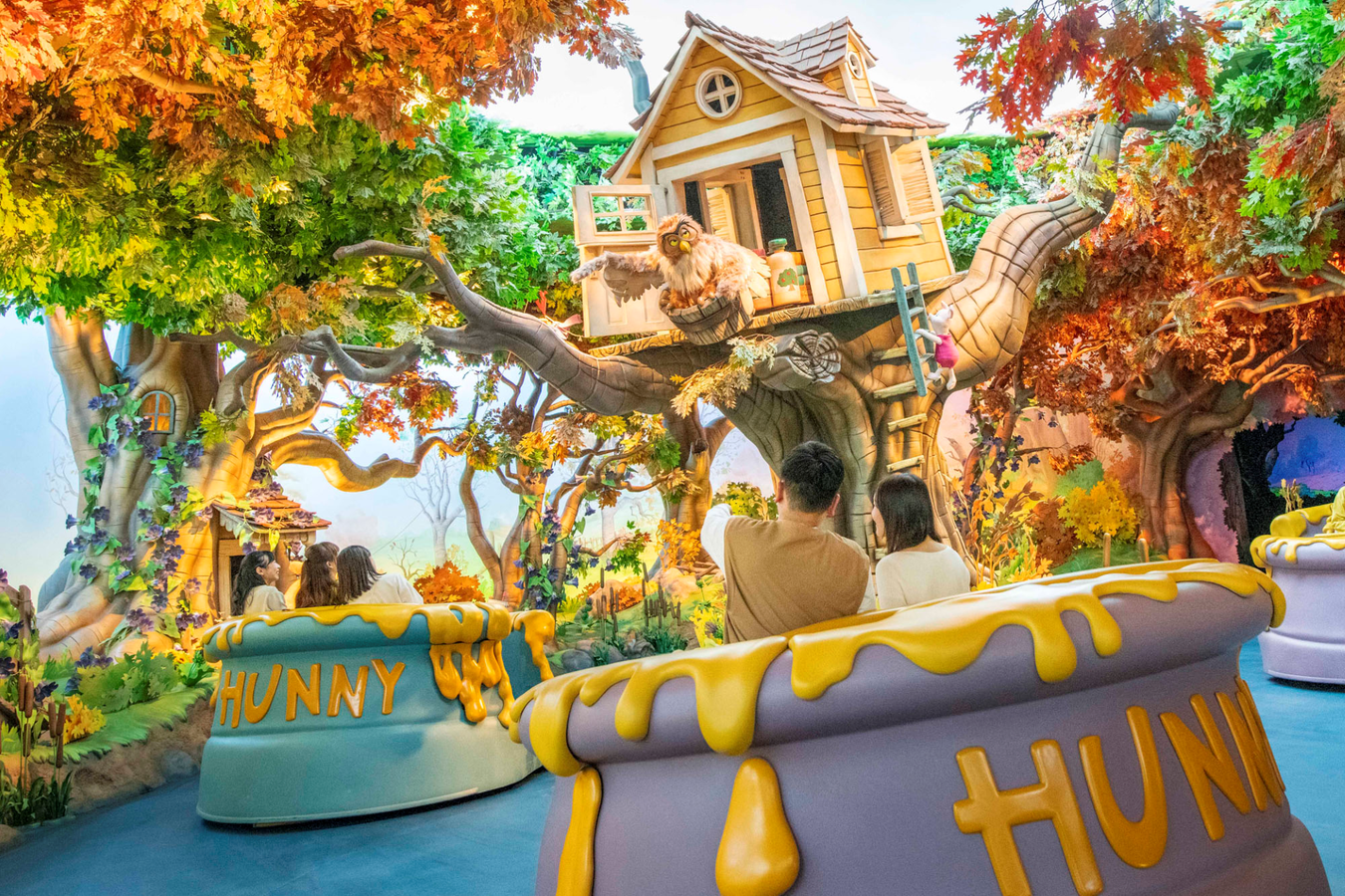 WinniethePoohHunnyHuntTokyo Fantasy Land News Whimsical treehouse ride on a large forked tree with autumn leaves and a lion figure near the doorway riders at the front A colorful HUNNY sign is visible on the ride base