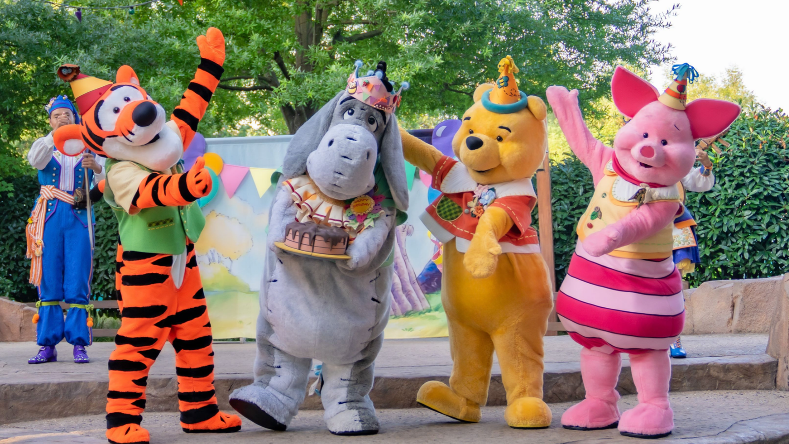 Costumed characters Tigger Eeyore Pooh and Piglet pose at a birthday party with a cake on a stage backdrop