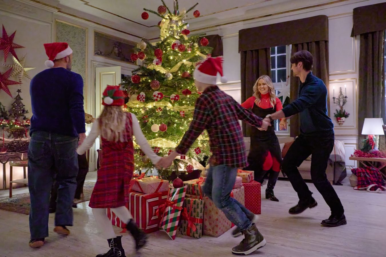 Group of people wearing Santa hats dancing in a circle around a lit Christmas tree with wrapped gifts in a cozy living room