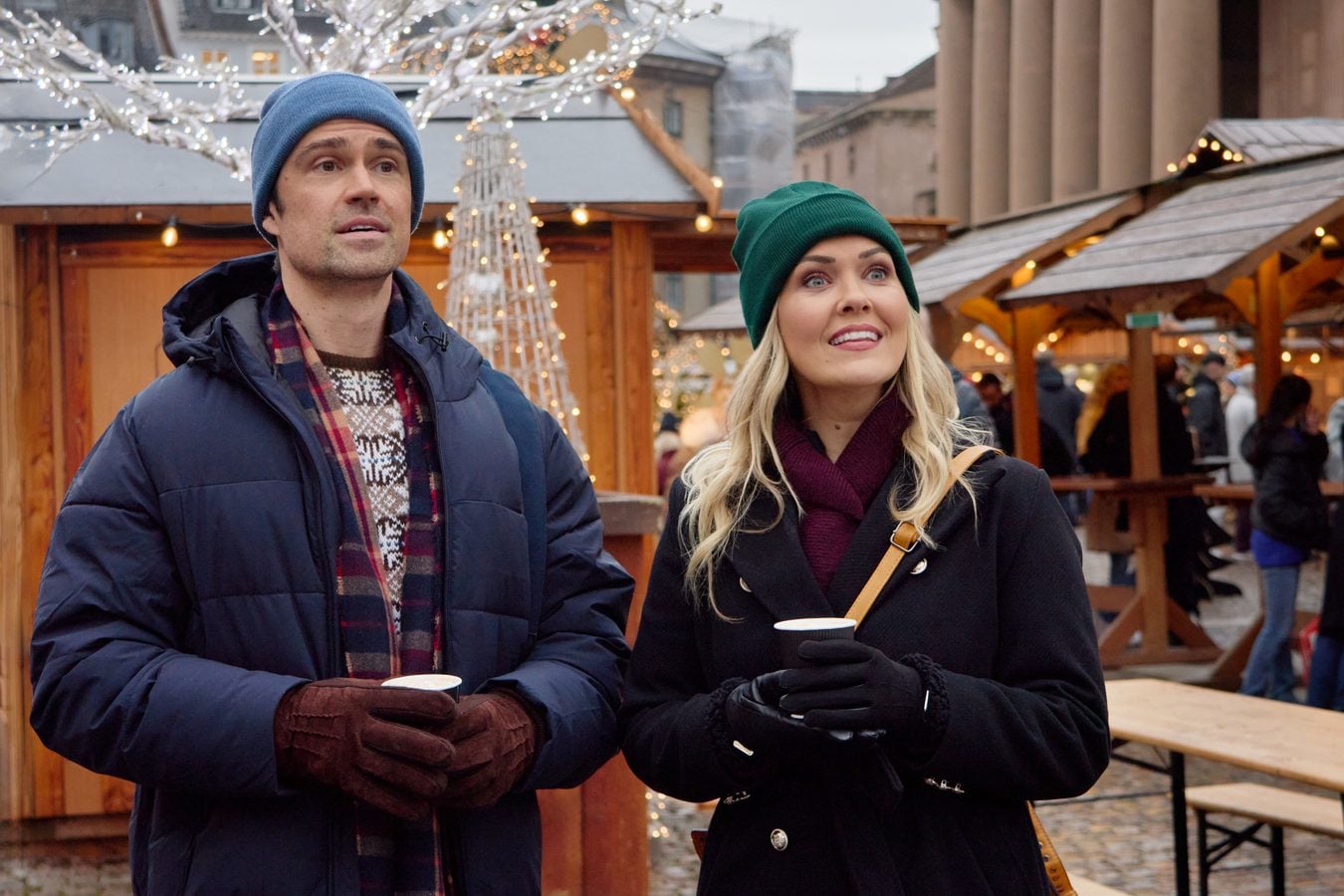 Two people in winter clothing stand at a Christmas market holding cups and smiling as lights glitter above wooden stalls