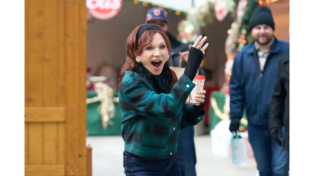 Woman in a teal plaid coat waves with a takeaway coffee in hand at a festive outdoor market