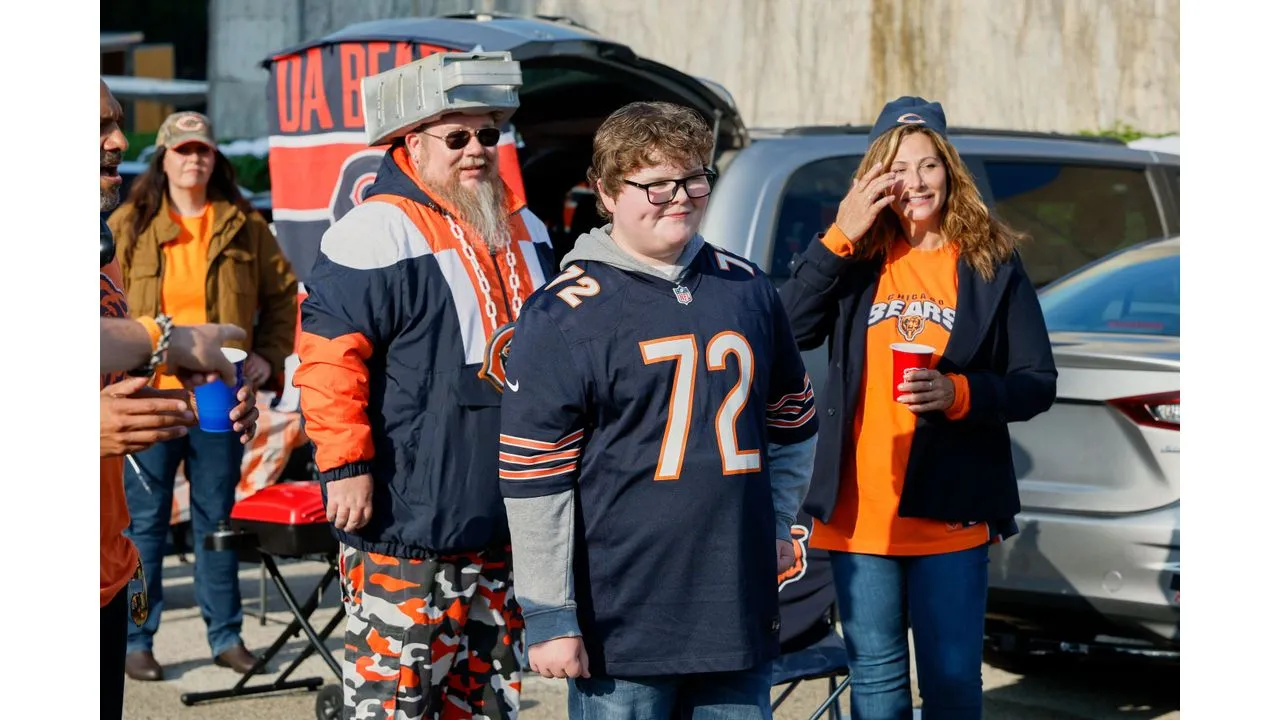 Group of Chicago Bears fans at a tailgate with a man in camouflage pants and a helmet a boy in a 72 Bears jersey and a woman in orange nearby near cars and a tent garden setup