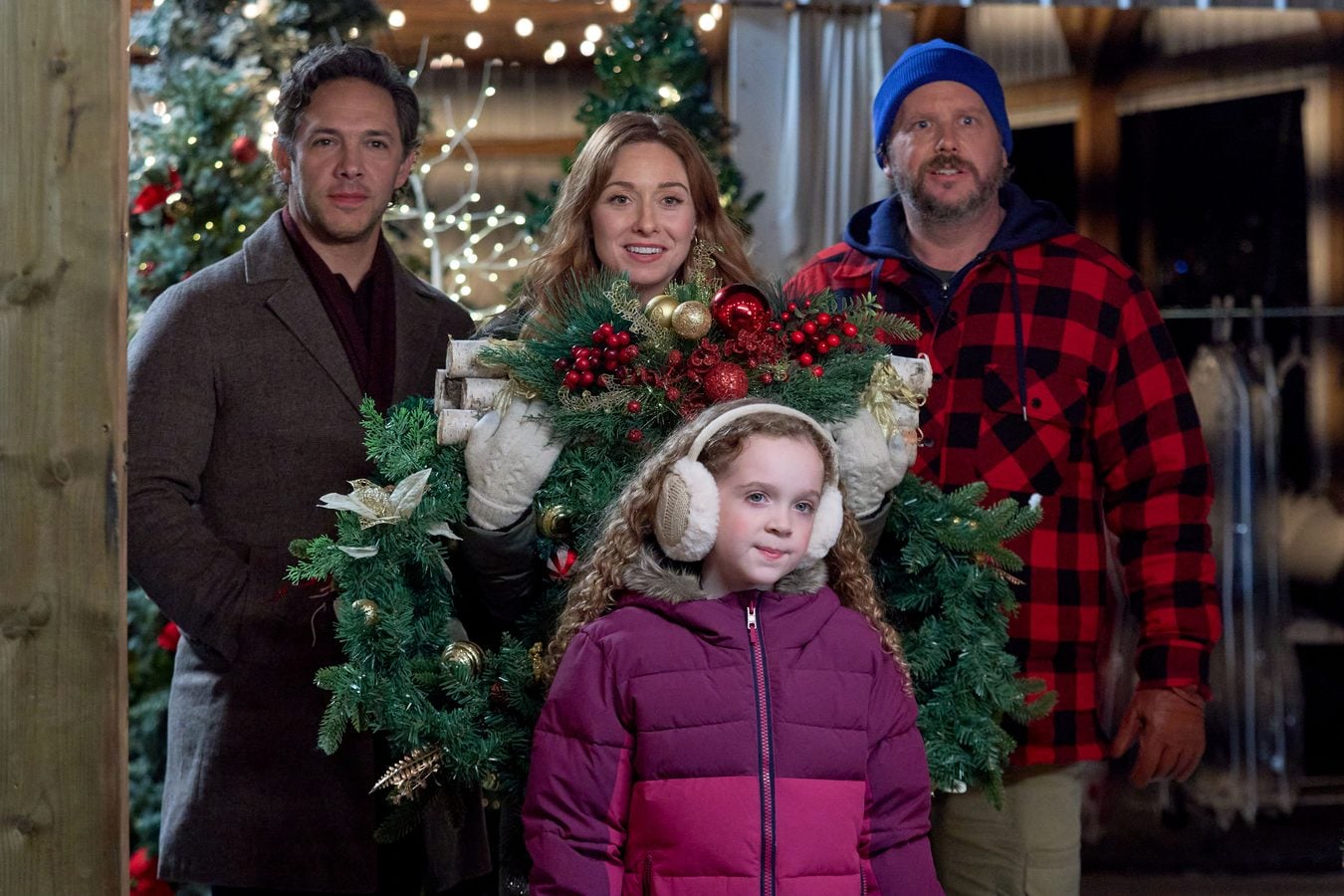 Amy Groening holds a large Christmas wreath behind Taylor Blake as Michael Rady and a co star in buffalo plaid look on in Adopting St Nick