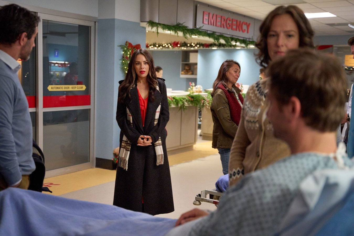 Woman in a black coat with a red top and plaid scarf stands in a hospital lobby near the emergency entrance Christmas garlands above