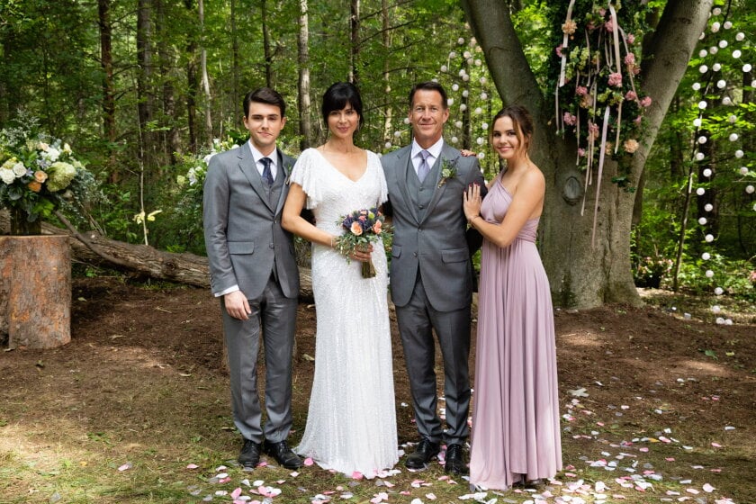 Bride in a white gown stands with two male guests in gray suits and a bridesmaid in a lavender dress outdoors with floral decor and petals on the ground at a forest wedding