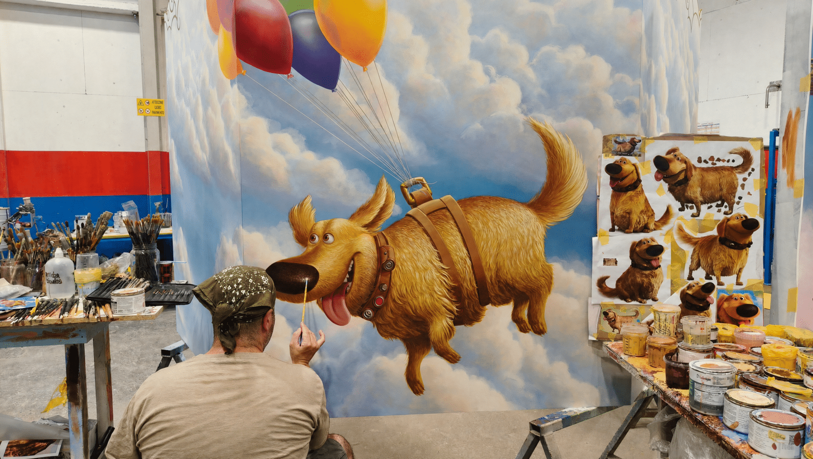 Painter adding details to a large golden retriever mural with a balloon filled sky art supplies and reference posters visible nearby