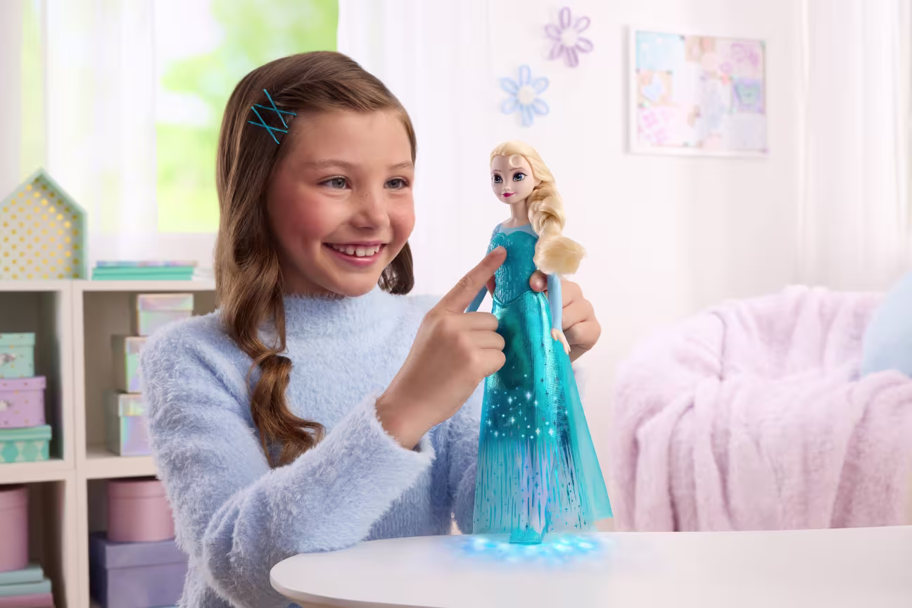 Smiling girl in a light blue sweater points at a glowing Elsa doll on a table in a pastel room