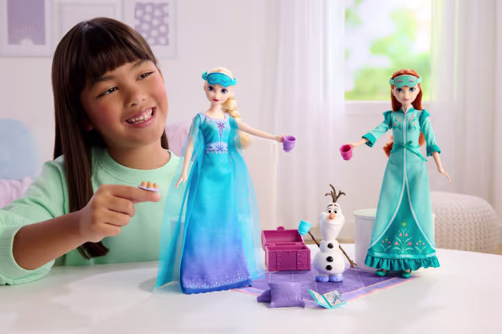 Smiling girl playing with blue Elsa and Anna dolls and a small Olaf figure on a white table in a bright room